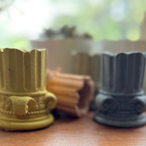 May include: Four small decorative planters made of concrete, each with a different color and design. The planters are shaped like columns with intricate details and are perfect for displaying small plants or succulents.