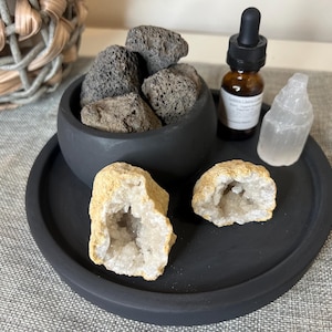 May include: A black tray with a black bowl filled with dark gray lava rocks. A clear glass bottle with a dropper and a white crystal are on the tray. Two white crystal geodes are on the tray.