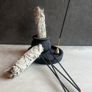 May include: A black incense holder with a gold-colored cone, several black incense sticks, and two bundles of white sage tied with string. The sage bundles are placed in a black pot-shaped holder.