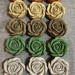 Puede incluir: Una variedad de decoraciones hechas a mano con forma de rosa en varios colores. Las rosas van desde tonos cálidos de la tierra como óxido y beige hasta tonos más fríos de verde y gris. Cada rosa está intrincadamente detallada.