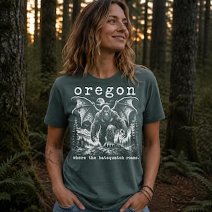 May include: A dark green short-sleeve t-shirt featuring a white graphic print of a winged creature in a forest setting. The text reads "oregon" at the top and "where the batsquatch roams." at the bottom, centred on the chest of the garment.