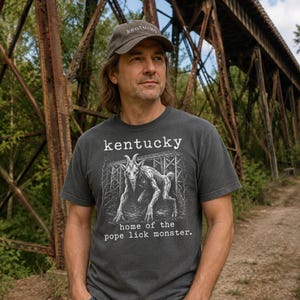 Op de afbeelding: Donkergrijs t-shirt met het woord "kentucky" boven een geitafbeelding en de tekst "home of the pope lick monster". Een bijpassende pet met "kentucky" erop. De persoon staat voor een roestige metalen brug.