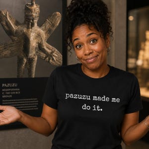 Puede incluir: Una camiseta negra con el texto blanco "pazuzu made me do it". La persona de la imagen sonríe y gesticula. Detrás hay una exhibición con una estatua de bronce y texto sobre Pazuzu.