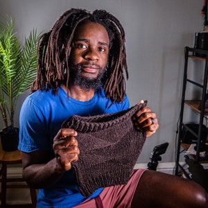 May include: A person with long dreadlocks is holding a brown knitted hat in front of them. The hat is made with a simple stitch pattern and has a ribbed brim.