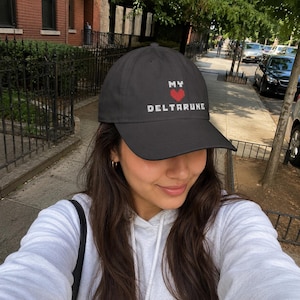 May include: A dark gray baseball cap featuring white pixelated text that reads MY and DELTARUNE, separated by a small red pixelated heart graphic. The cap has a curved brim and a structured crown, displaying a clean and simple design aesthetic.