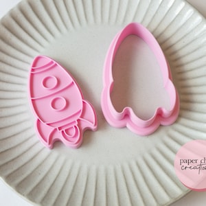 May include: Two pink plastic cookie cutters shaped like a rocket ship. The cutters are on a white plate with a textured surface. The text "paper chain creations" is on a pink circle sticker in the lower right corner of the image.