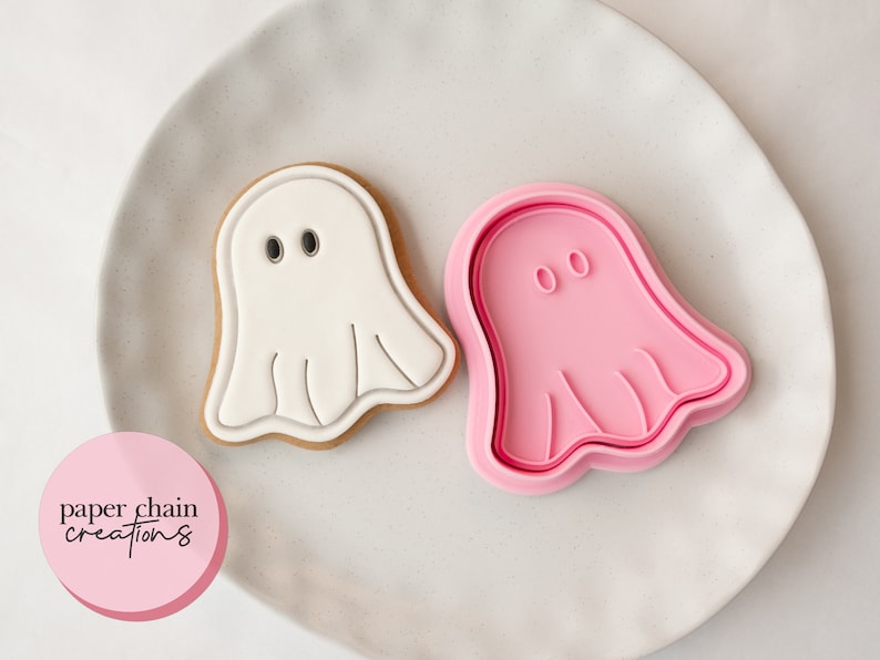 May include: A white ghost-shaped cookie with black eyes and a pink ghost-shaped cookie cutter. The cookie is on a white plate. The cookie cutter is pink. The image includes the text "paper chain creations" in a pink circle.