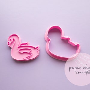 May include: Two pink plastic cookie cutters, one shaped like a swan and the other shaped like a duck. The cutters are on a light purple background. The text "paper chain creations" is visible in the lower right corner of the image.