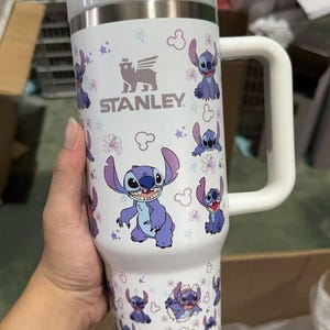 Op de afbeelding: Witte Stanley-beker met Stitch-ontwerp. De beker heeft een roestvrijstalen bovenkant en een handvat. Het ontwerp bevat Stitch-illustraties, bloemen en het Stanley-logo.