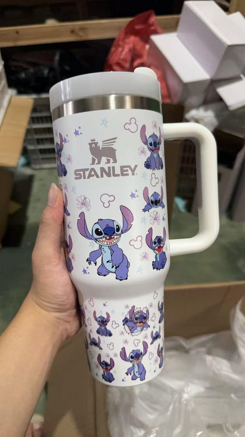 Op de afbeelding: Witte Stanley-beker met Stitch-ontwerp. De beker heeft een roestvrijstalen bovenkant en een handvat. Het ontwerp bevat Stitch-illustraties, bloemen en het Stanley-logo.