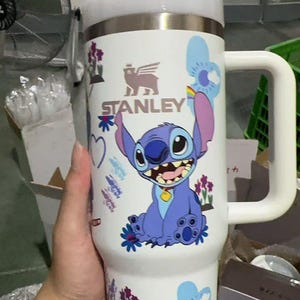 May include: A white Stanley tumbler featuring a cartoon Stitch design. The tumbler has a large handle, a white lid, and a stainless steel body. The design includes Stitch, flowers, and the Stanley logo.