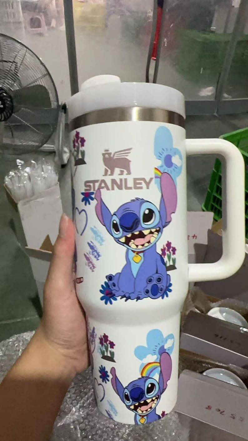 May include: A white Stanley tumbler featuring a cartoon Stitch design. The tumbler has a large handle, a white lid, and a stainless steel body. The design includes Stitch, flowers, and the Stanley logo.