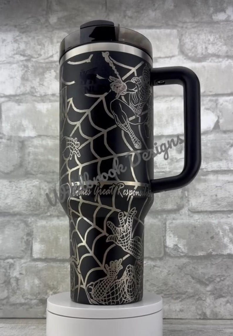 Puede incluir: Vaso negro con un dise&ntilde;o plateado de Spider-Man, que incluye un patr&oacute;n de telara&ntilde;a y el superh&eacute;roe. El vaso tiene un asa y tapa negras, y el texto "With Great Responsibility" es visible.
