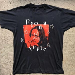 K&ouml;nnte beinhalten: Schwarzes T-Shirt mit rotem Quadrat, das ein Frauenportr&auml;t und den Namen "Fiona Apple" in Wei&szlig; zeigt. Wei&szlig;e Sterne umgeben das rote Quadrat. Ein Vintage-Artikel zum Thema Musik.