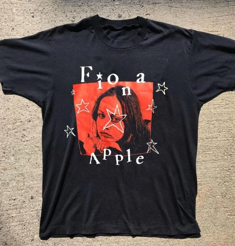 K&ouml;nnte beinhalten: Schwarzes T-Shirt mit rotem Quadrat, das ein Frauenportr&auml;t und den Namen "Fiona Apple" in Wei&szlig; zeigt. Wei&szlig;e Sterne umgeben das rote Quadrat. Ein Vintage-Artikel zum Thema Musik.