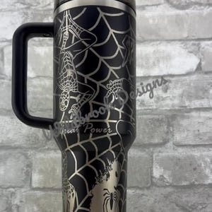 Puede incluir: Vaso t&eacute;rmico negro con asa, con un dise&ntilde;o de telara&ntilde;a plateada e ilustraciones de Spider-Man. Tambi&eacute;n es visible el texto "With Great Power". Incluye tapa y pajita negras.