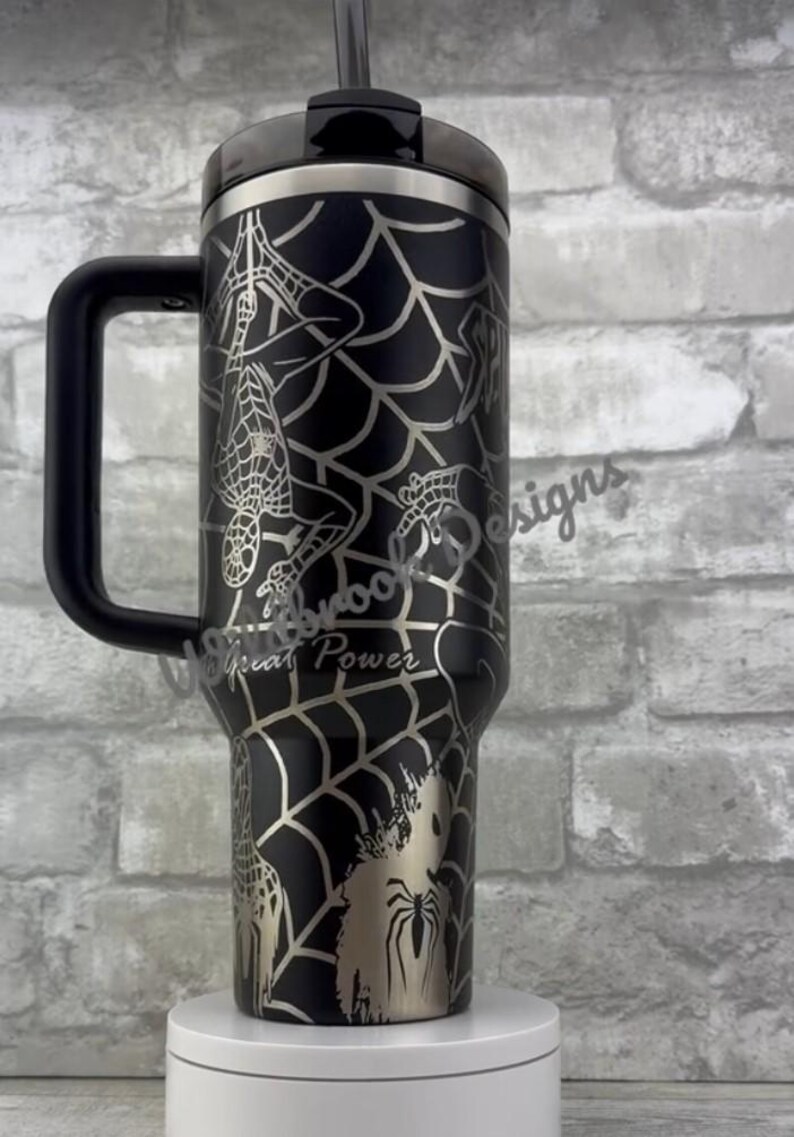 Puede incluir: Vaso t&eacute;rmico negro con asa, con un dise&ntilde;o de telara&ntilde;a plateada e ilustraciones de Spider-Man. Tambi&eacute;n es visible el texto "With Great Power". Incluye tapa y pajita negras.