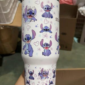 Op de afbeelding: Een witte beker met een herhaald patroon van het Disney-personage Stitch. Het ontwerp toont Stitch in verschillende poses, omringd door sterren en bloemen. De beker heeft een zilveren deksel.