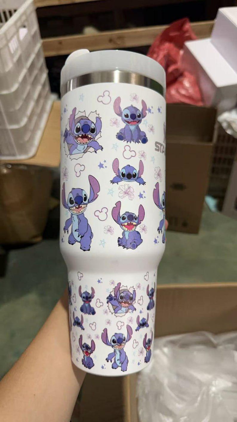 Op de afbeelding: Een witte beker met een herhaald patroon van het Disney-personage Stitch. Het ontwerp toont Stitch in verschillende poses, omringd door sterren en bloemen. De beker heeft een zilveren deksel.