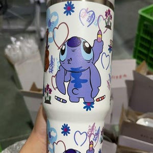 May include: White insulated tumbler featuring a cartoon character in shades of purple. The tumbler is decorated with hearts, flowers, and musical instruments. The tumbler has a white lid and is designed to keep beverages cold or hot.