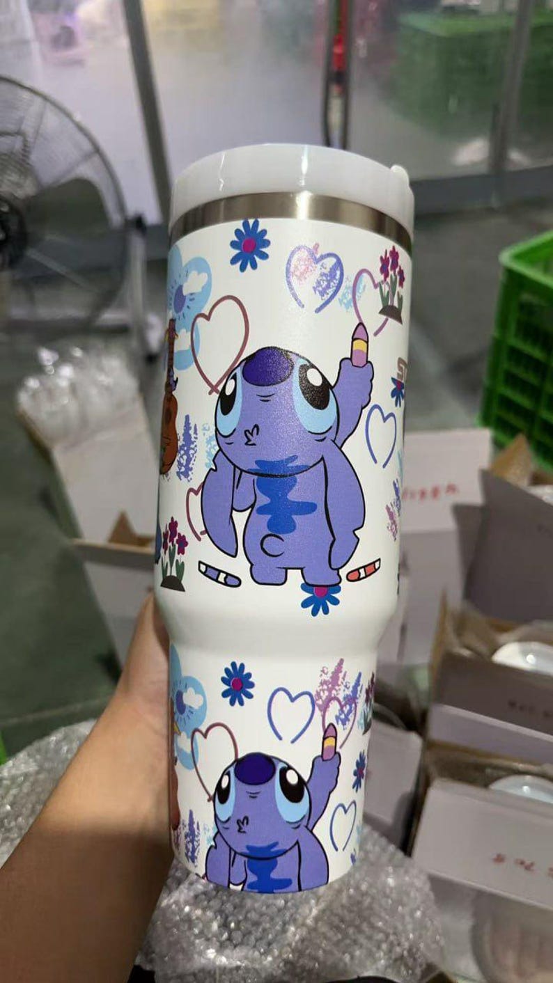 May include: White insulated tumbler featuring a cartoon character in shades of purple. The tumbler is decorated with hearts, flowers, and musical instruments. The tumbler has a white lid and is designed to keep beverages cold or hot.