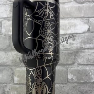 Puede incluir: Vaso negro con un dise&ntilde;o plateado de Spider-Man, que incluye un patr&oacute;n de telara&ntilde;a y la silueta del personaje. El vaso tiene un asa negra y un borde plateado. El texto "With Great Power" es visible.