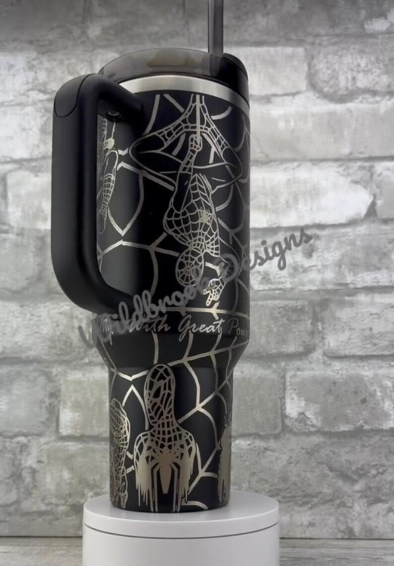 Puede incluir: Vaso negro con un dise&ntilde;o plateado de Spider-Man, que incluye un patr&oacute;n de telara&ntilde;a y la silueta del personaje. El vaso tiene un asa negra y un borde plateado. El texto "With Great Power" es visible.