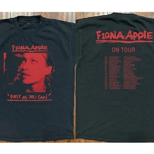 Op de afbeelding: Zwart t-shirt met een rode afbeelding van Fiona Apple en de tekst "Fast As You Can" op de voorkant. De achterkant van het shirt toont "Fiona Apple On Tour" in het rood, samen met een lijst met tourdata en locaties.