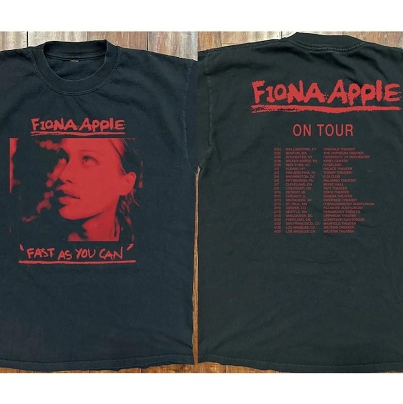 Op de afbeelding: Zwart t-shirt met een rode afbeelding van Fiona Apple en de tekst "Fast As You Can" op de voorkant. De achterkant van het shirt toont "Fiona Apple On Tour" in het rood, samen met een lijst met tourdata en locaties.