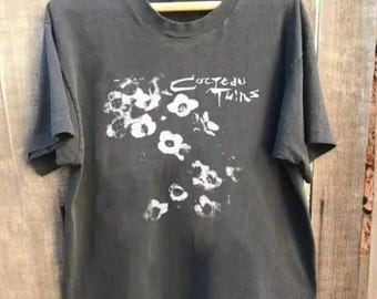 May include: Dark gray t-shirt with a white floral design and the band name "Cocteau Twins" printed above the flowers. The shirt is hanging on a wooden hanger against a wooden background.