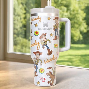 May include: A white Stanley tumbler featuring a Woody design from Toy Story. The tumbler has a white handle and lid, with illustrations of Woody, stars, and the word "Woody" in a playful font. The Stanley logo is also visible.