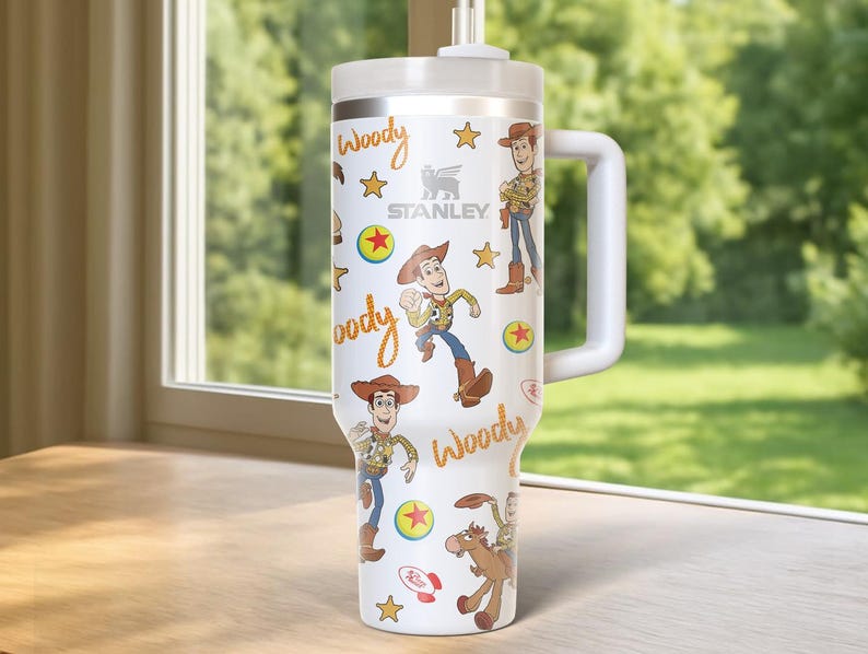 May include: A white Stanley tumbler featuring a Woody design from Toy Story. The tumbler has a white handle and lid, with illustrations of Woody, stars, and the word "Woody" in a playful font. The Stanley logo is also visible.