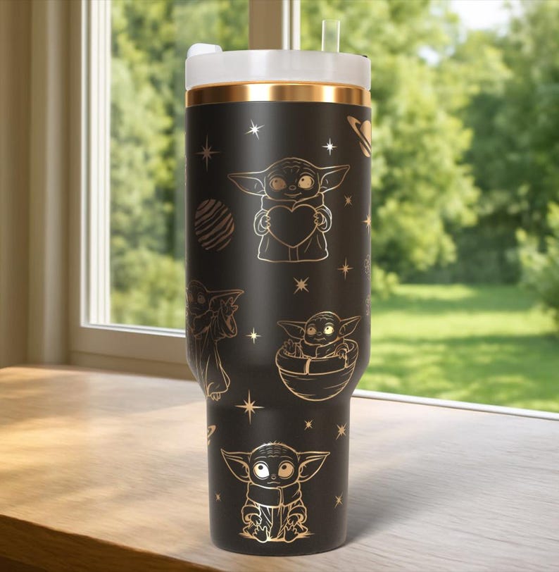 Puede incluir: Vaso negro con detalles dorados y tapa blanca. El vaso presenta ilustraciones doradas de un personaje sosteniendo un coraz&oacute;n, en una c&aacute;psula y otros dise&ntilde;os. El vaso tiene una banda dorada cerca de la parte superior y una pajita.