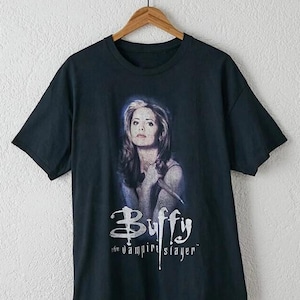 May include: A black short-sleeve t-shirt featuring a graphic print of a person with long hair holding a wooden stake. Below the image, the text reads "Buffy the vampire slayer" in a stylized white font. The shirt is displayed on a wooden hanger.