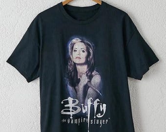 May include: A black short-sleeved t-shirt featuring a graphic print of a person with long hair holding a wooden stake. Below the image, the text reads "Buffy the vampire slayer" in a stylised white font. The shirt is displayed on a wooden hanger.