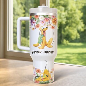 Może przedstawiać: Biały kubek z kresk&oacute;wkowym wizerunkiem psa Pluto, otoczony kolorowymi kwiatami. Kubek ma uchwyt i pokrywkę. Na kubku wydrukowano napis "YOUR NAME". Projekt zawiera zabawną ilustrację Pluto.