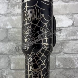 Puede incluir: Vaso de acero inoxidable negro con un dise&ntilde;o de Spider-Man. El vaso tiene un patr&oacute;n de telara&ntilde;a plateada y las palabras "Spider-Man" en una fuente estilizada. El dise&ntilde;o incluye una imagen de Spider-Man y una ara&ntilde;a.