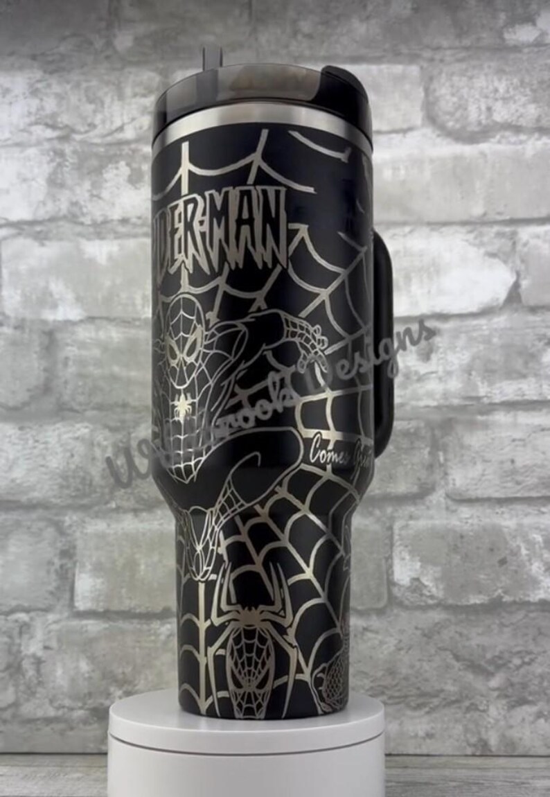 Puede incluir: Vaso de acero inoxidable negro con un dise&ntilde;o de Spider-Man. El vaso tiene un patr&oacute;n de telara&ntilde;a plateada y las palabras "Spider-Man" en una fuente estilizada. El dise&ntilde;o incluye una imagen de Spider-Man y una ara&ntilde;a.
