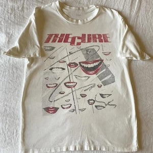 May include: Off-white t-shirt featuring the band name "THE CURE" in red block letters. The shirt's graphic design includes multiple red lips and gray line art. The t-shirt is made of a soft, comfortable material.