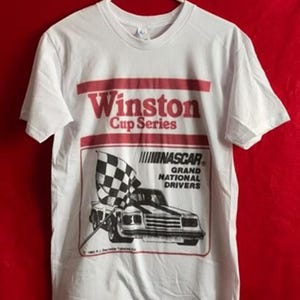 Puede incluir: Una camiseta blanca de manga corta con un estampado gr&aacute;fico en rojo y negro. El texto dice Winston Cup Series, NASCAR y Grand National Drivers. Una bandera a cuadros blanca y negra y un coche de carreras aparecen dentro de un marco rojo.