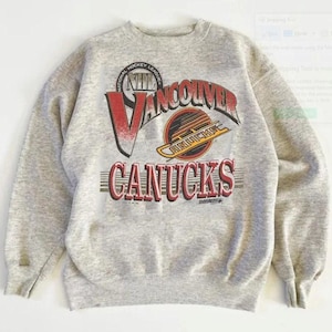 Pu&ograve; includere: Felpa girocollo grigio chiaro con il logo dei Vancouver Canucks. Il logo include il nome della squadra in rosso e bianco, con un contorno nero, e un emblema circolare con il logo NHL. Articolo vintage.
