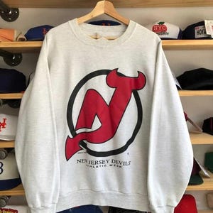 May include: A light gray sweatshirt featuring the New Jersey Devils logo in red and black. The logo is a stylized "N" with horns inside a black circle. The text "NEW JERSEY DEVILS ATHLETIC WEAR" is printed below the logo.