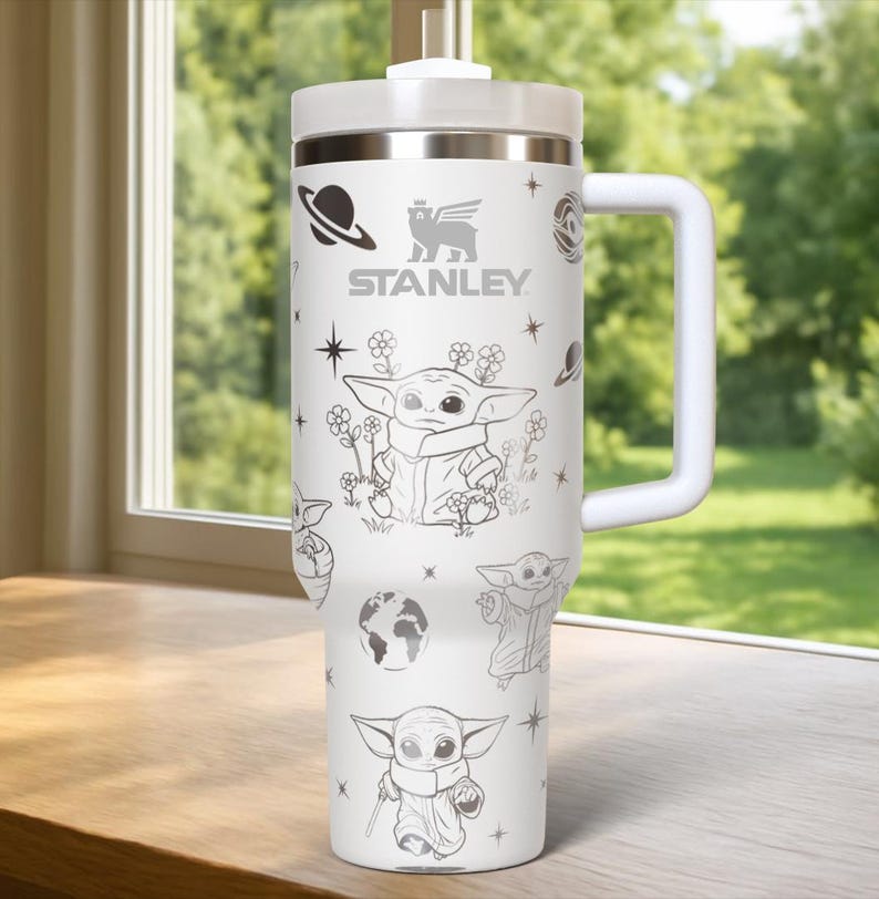 Puede incluir: Vaso t&eacute;rmico blanco con asa, con un dise&ntilde;o de Baby Yoda y gr&aacute;ficos espaciales. El vaso tiene el logotipo "STANLEY" y una tapa blanca. El dise&ntilde;o incluye ilustraciones de planetas, estrellas y Baby Yoda en varias poses.
