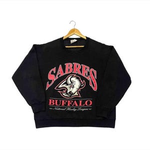 May include: Black sweatshirt with the word "SABRES" in red arched lettering above a white and gray buffalo head logo. Below the logo, the word "BUFFALO" is in red, with "National Hockey League" underneath.