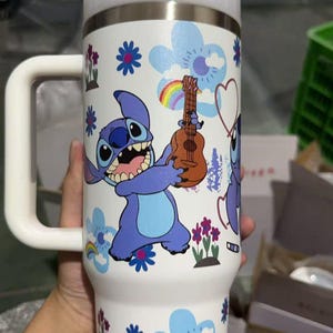May include: White insulated tumbler featuring a cartoon character holding a ukulele. The tumbler has a white handle and lid, with a design of blue flowers, rainbows, and hearts. The character is blue with large eyes and a wide grin.