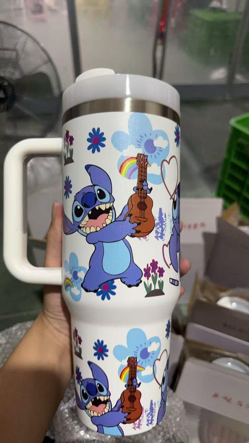 May include: White insulated tumbler featuring a cartoon character holding a ukulele. The tumbler has a white handle and lid, with a design of blue flowers, rainbows, and hearts. The character is blue with large eyes and a wide grin.