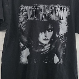May include: Black t-shirt with a grayscale image of Siouxsie Sioux and the band name, Siouxsie and the Banshees, printed above. The graphic is distressed, in white. The shirt has a crew neck and short sleeves.