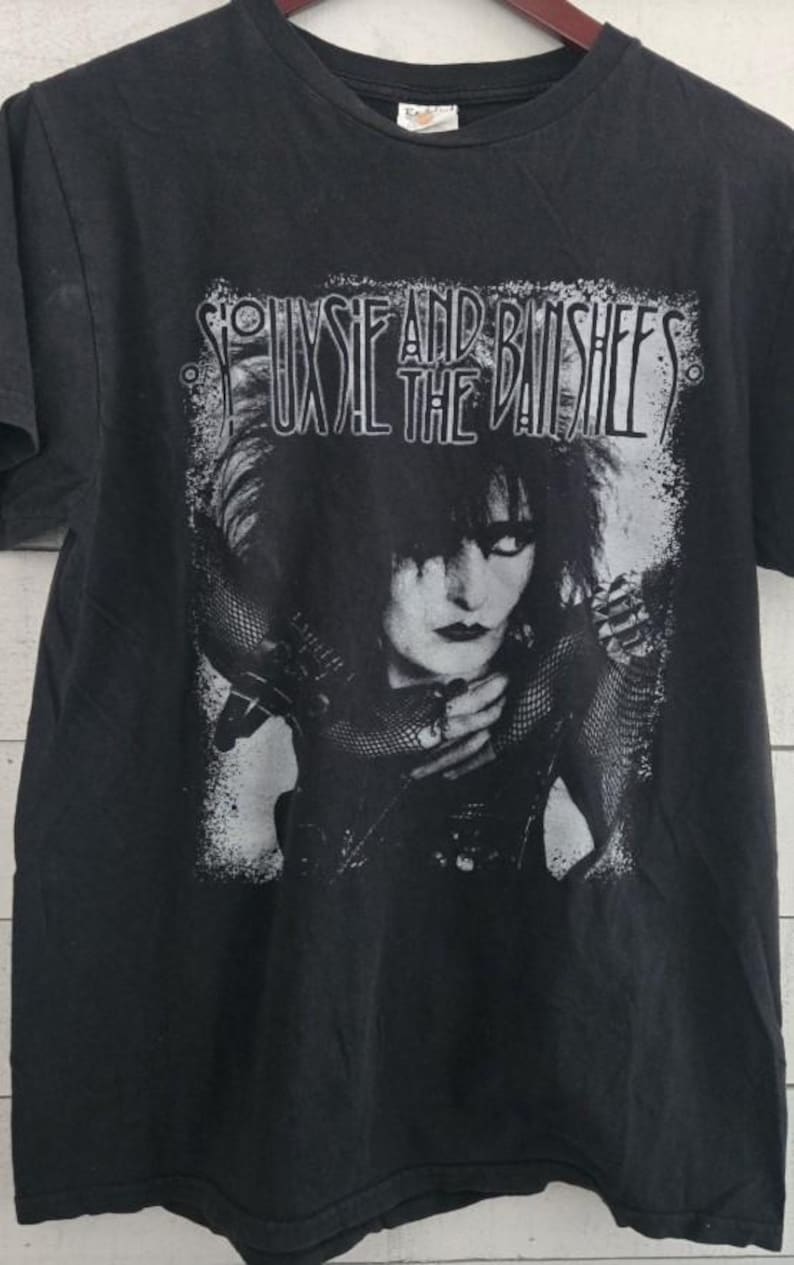 May include: Black t-shirt with a grayscale image of Siouxsie Sioux and the band name, Siouxsie and the Banshees, printed above. The graphic is distressed, in white. The shirt has a crew neck and short sleeves.