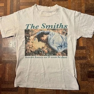 May include: White t-shirt featuring the band name "The Smiths" in teal lettering above a photo of a person lying down. The text "heaven knows we'll soon be dust" is below the photo. The shirt has short sleeves and a crew neck.