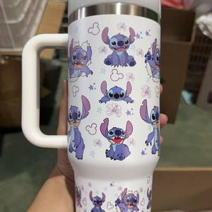 Op de afbeelding: Witte ge&iuml;soleerde beker met een herhaald patroon van het personage Stitch uit Lilo & Stitch. De beker heeft een witte handgreep en deksel. Het ontwerp bevat Stitch, bloemen en sterren. Geschikt voor warme en koude dranken.